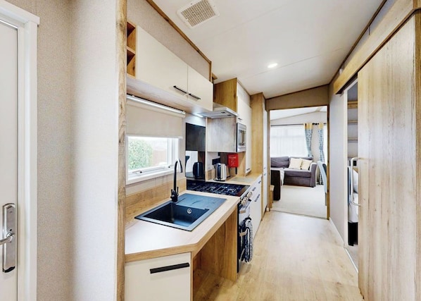 A compact kitchenette with a sink, stove, and cabinets, leading to a living area with a couch and window.