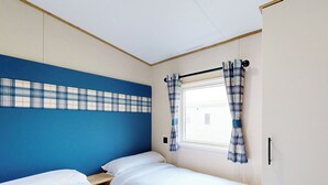 A room with two beds, a window with curtains, a blue wall, and a wooden headboard. - Gold 3 Bedroom Holiday Home Pet (Hornsea)