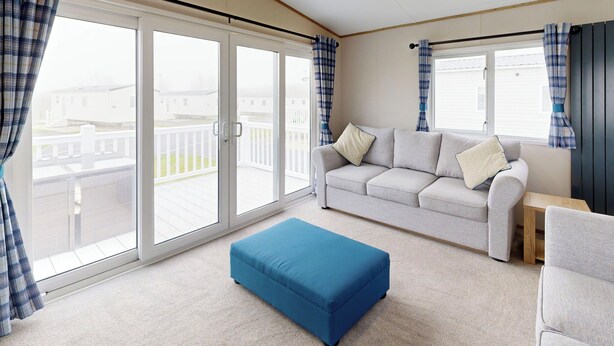 A room with a grey sofa, a blue ottoman, and a window with curtains. - Gold 3 Bedroom Holiday Home Pet (Hornsea)