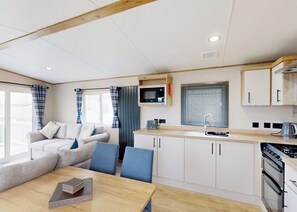 A modern kitchen with a microwave, stove, and sink. There is a dining area with a table and chairs. - Gold 3 Bedroom Holiday Home Pet (Hornsea)