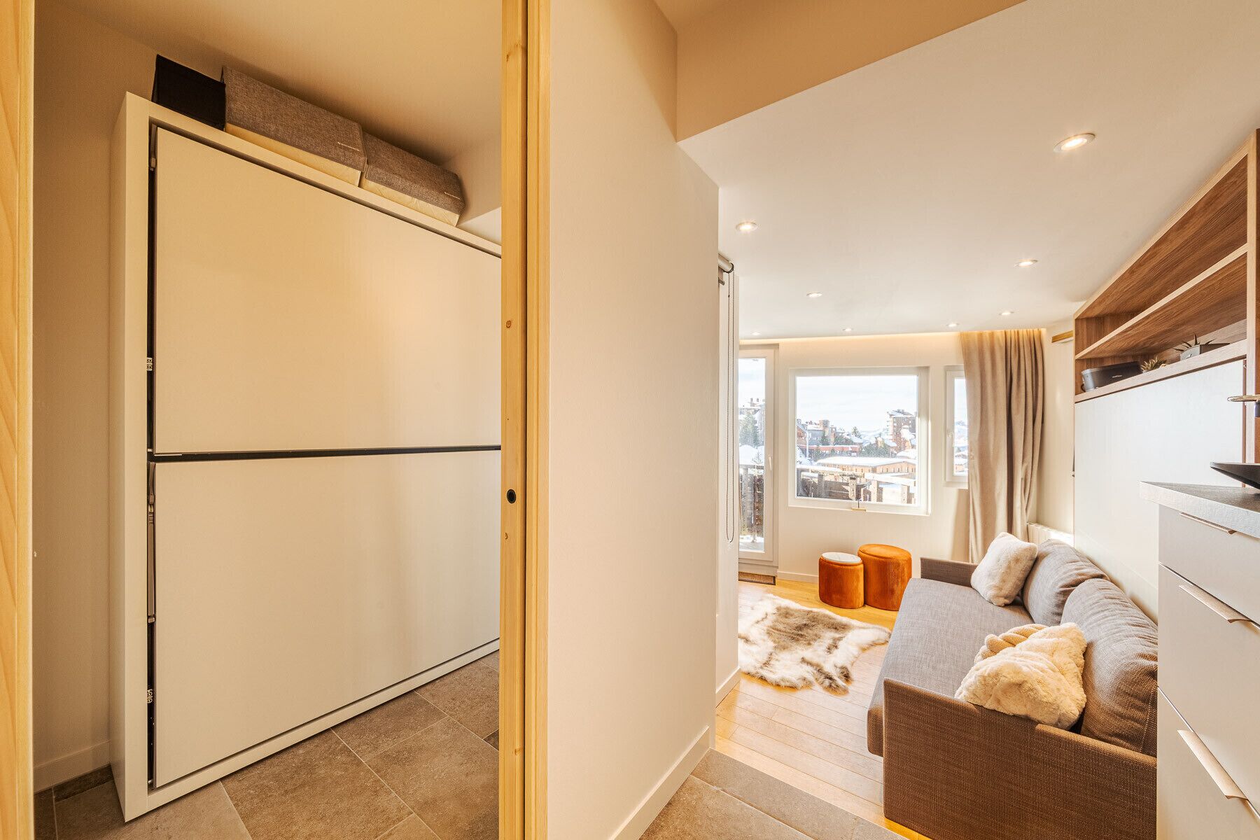 A modern apartment with a large wardrobe, a cozy seating area with a sofa and cushions, and a view of the city through a large window.