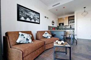 A modern living room with a sofa and a coffee table, a kitchen area with bar stools, and a framed cityscape picture on the wall. - Delizioso monolocale con ampio terrazzo a Testaccio (ROMA)