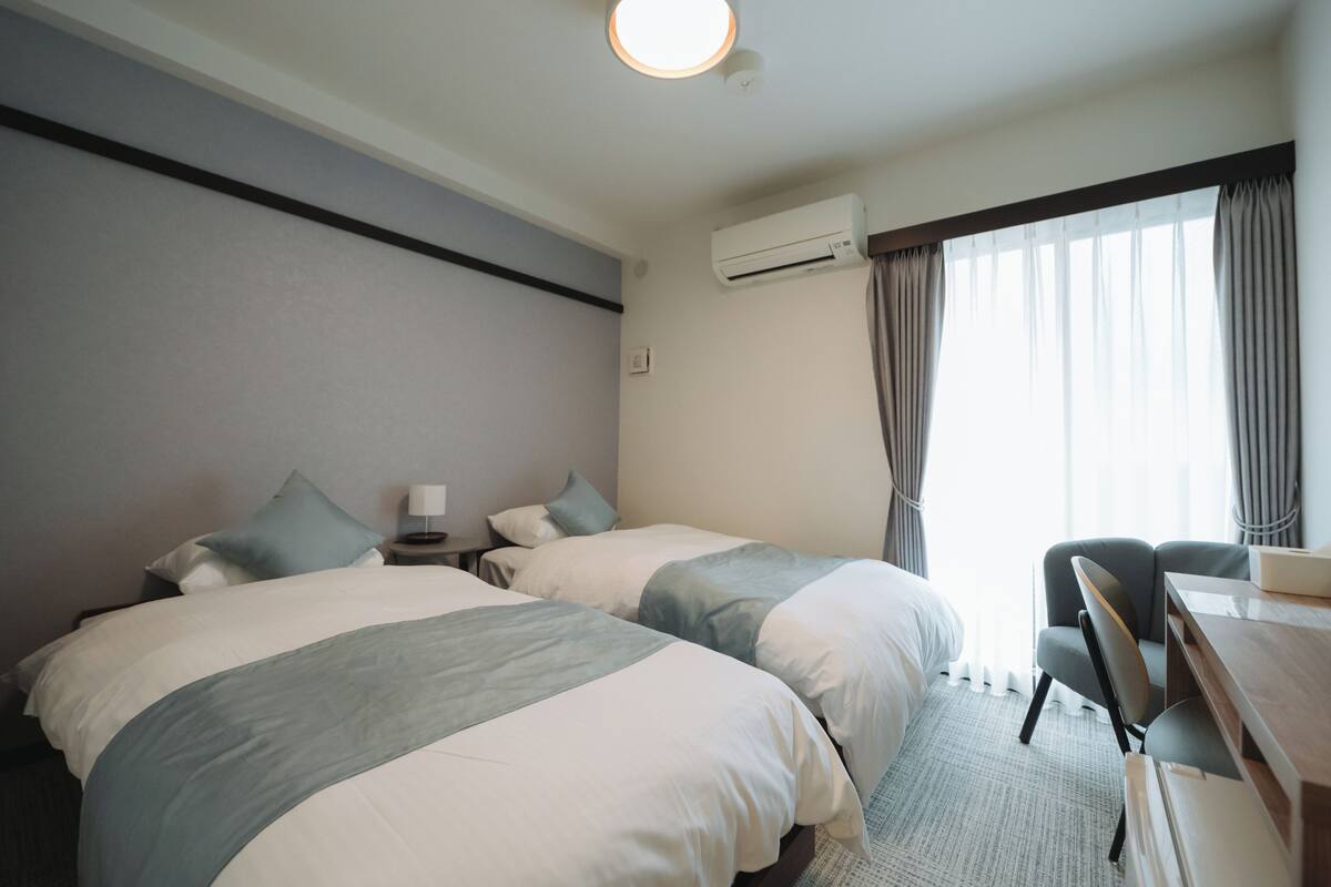 A hotel room with two beds, a desk, a chair, an air conditioning unit, and a window with curtains.