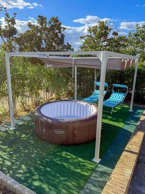 Ein Garten mit einem runden Whirlpool, zwei blau gestreiften Liegestühlen und einer Pergola.