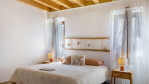 Un dormitorio con techo de madera, una cama con cubrecama floral, dos mesitas de noche con lámparas y un estante en la pared con adornos.