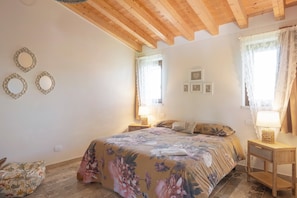 A bedroom with a floral bedspread, wooden beams, two bedside tables with lamps, and a window with curtains. - Double Room '4' with Shared Pool, Wi-Fi and Air Conditioning (Cavaion Veronese)