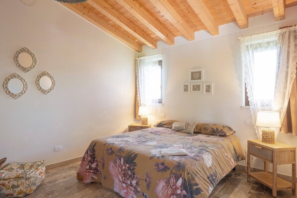 A bedroom with a floral bedspread, wooden beams, two bedside tables with lamps, and a window with curtains. - Double Room '4' with Shared Pool, Wi-Fi and Air Conditioning (Cavaion Veronese)