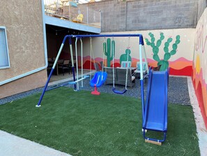 Ein Spielplatz mit Schaukeln und einer Rutsche, umgeben von Kunstgrün und einer Wandmalerei.