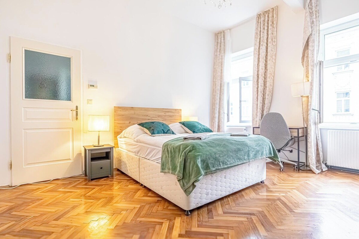 A bedroom with a bed, bedside table, lamp, chair, and large window with curtains.