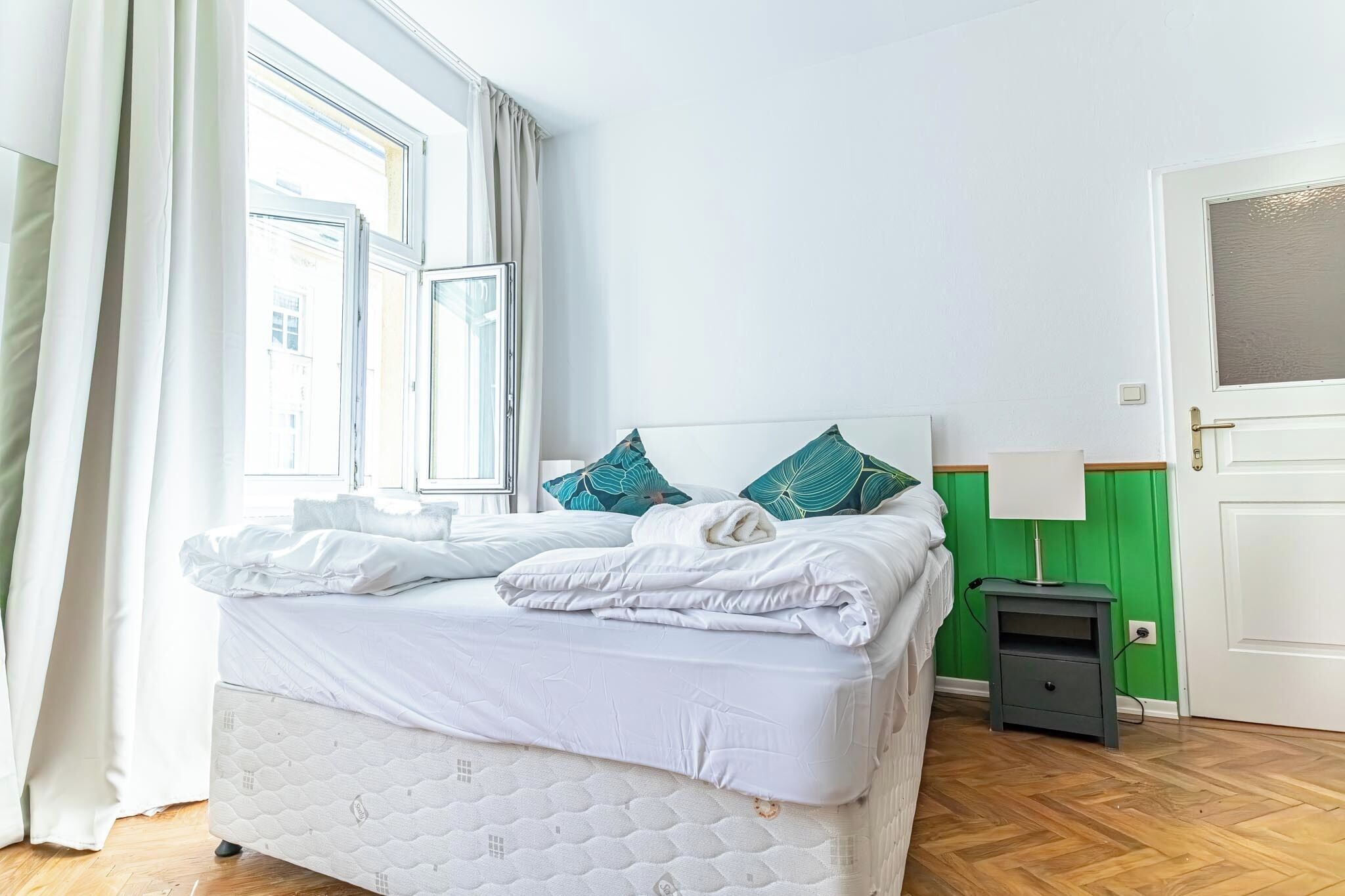 Una cama bien hecha con ropa blanca, una pared verde y una mesita de noche con una lámpara.