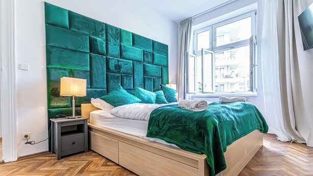 A modern bedroom with a large green headboard, a wooden bed frame, a nightstand with a lamp, and a window with a view of buildings. - Cozy Room Near Landstraße for Long Stay (Vienna)