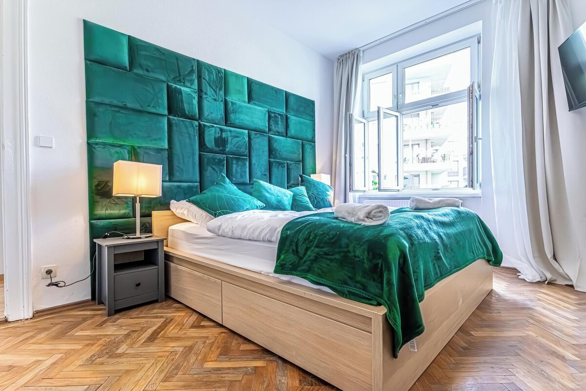 A modern bedroom with a large green headboard, a wooden bed frame, a nightstand with a lamp, and a window with a view of buildings.