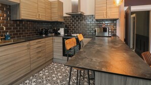 A modern kitchen with wooden cabinets, a black tiled backsplash, a stainless steel range hood, and a large island with chairs. - The Hyde (Preston)