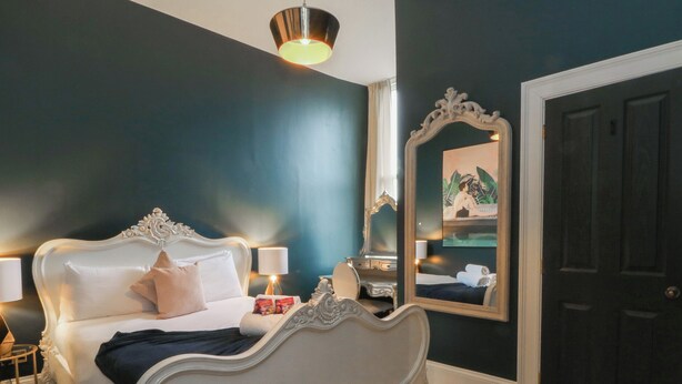 A bedroom with a white ornate bed, a bedside table with a lamp, a mirror, and a dark door. - The Hyde (Preston)