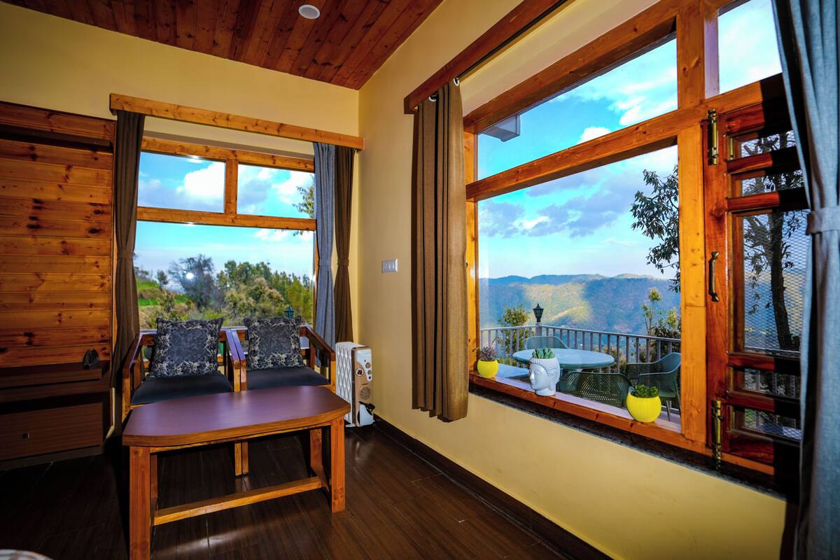 A wooden interior with a table and chairs, large windows with curtains, and a view of the outdoors.