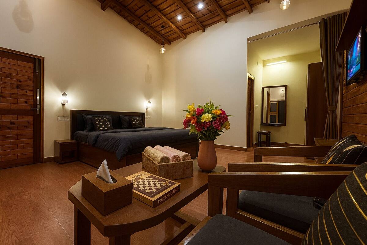 A cozy hotel room with a wooden ceiling, a bed with dark bedding, a bedside table with a tissue box and a chessboard, a vase with flowers, and a television on the wall.