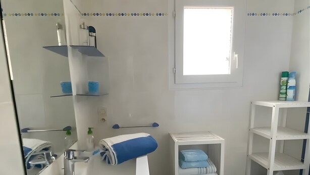 Et badeværelse med en hvid vask, spejl og hylder, et vindue og et toilet.