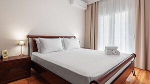 Ein ordentlich bezogenes Bett mit weißen Leinen, einem Holz-Kopfbrett und einem Nachttisch mit Lampe und Bilderrahmen.