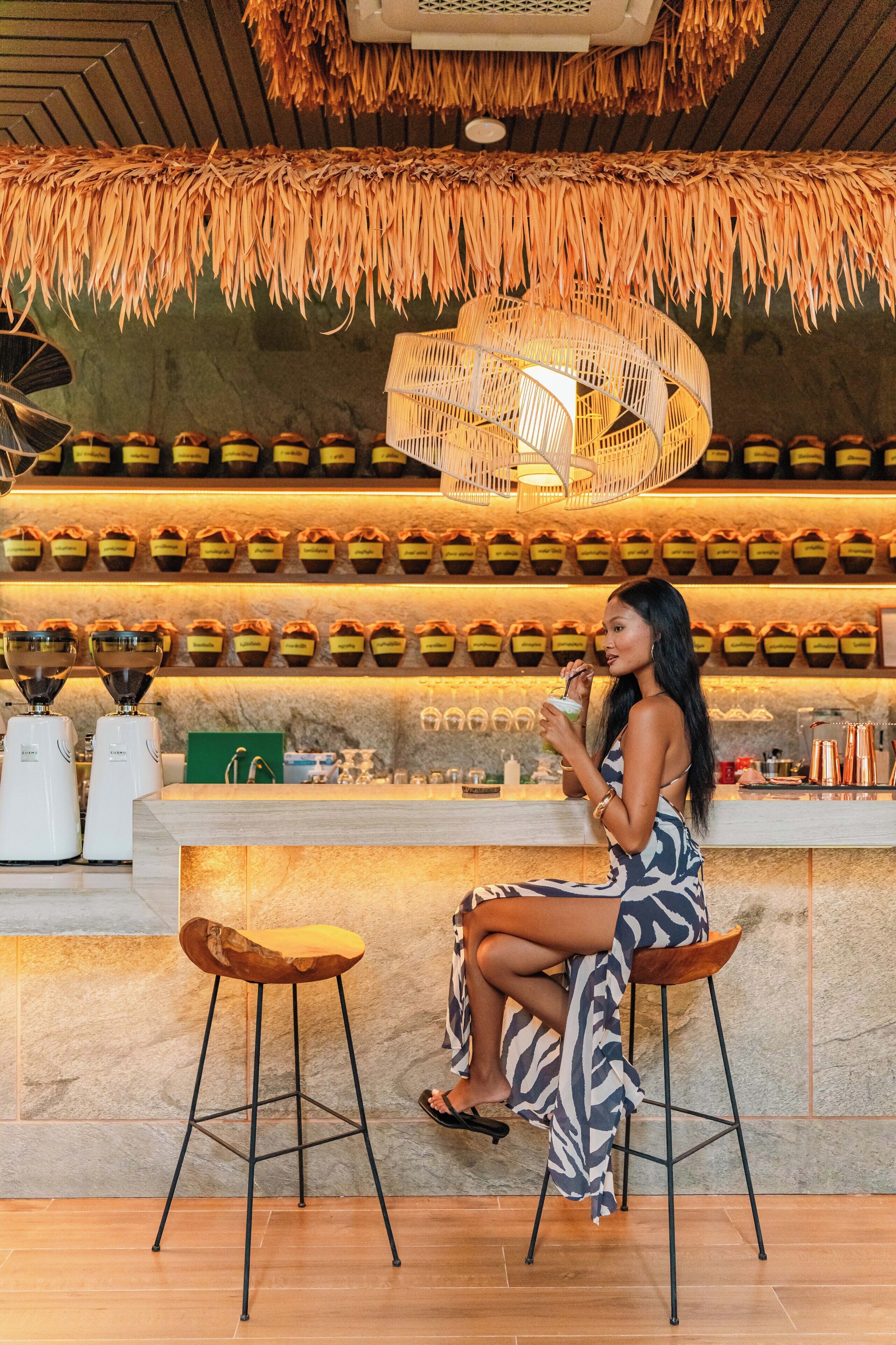 A person seated at a bar with a thatched ceiling, a counter with jars, and a wicker light fixture.