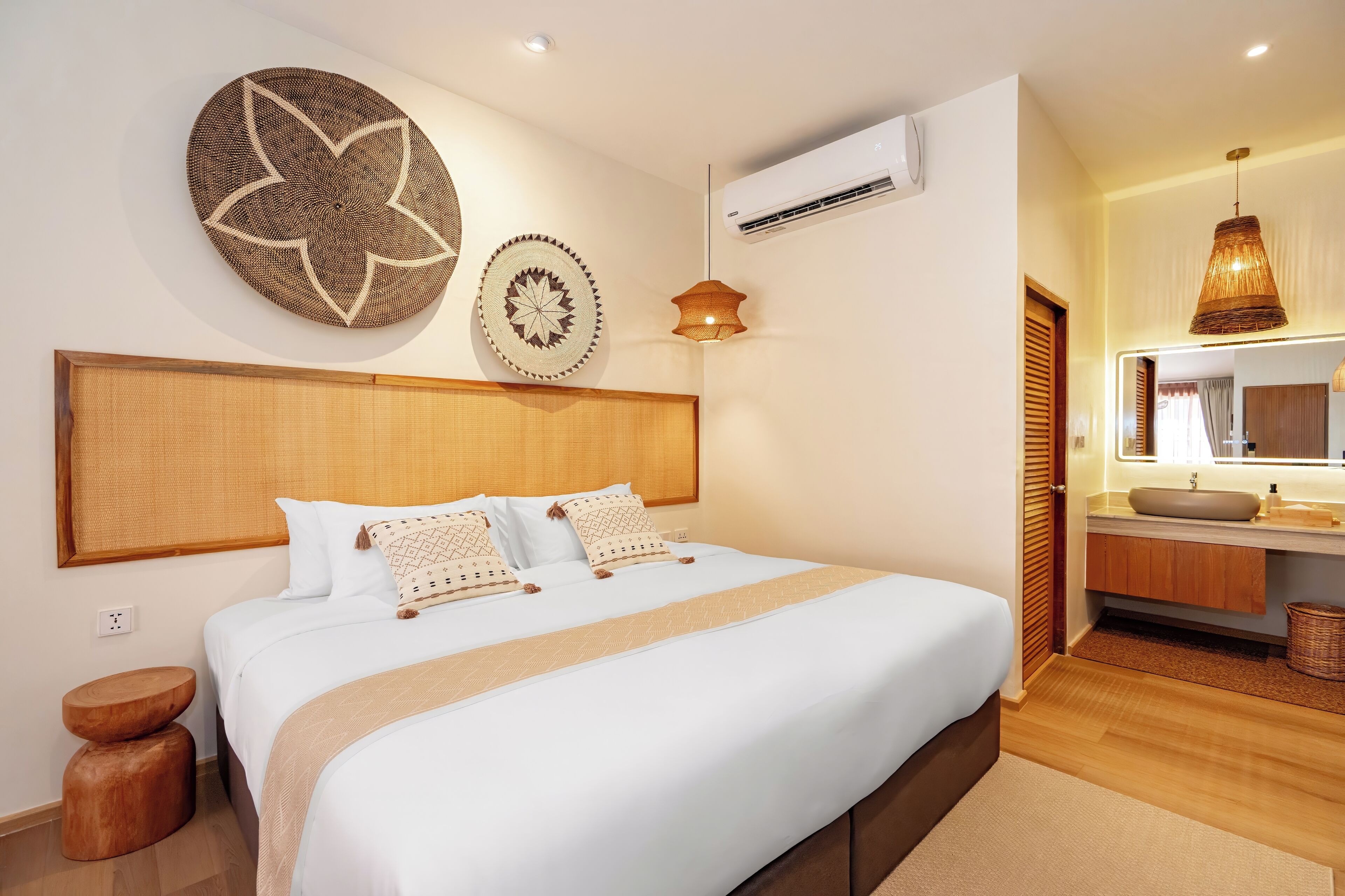 A hotel room with a large bed, a wooden headboard, a round wall decoration, a hanging light fixture, and a bathroom with a mirror and sink.