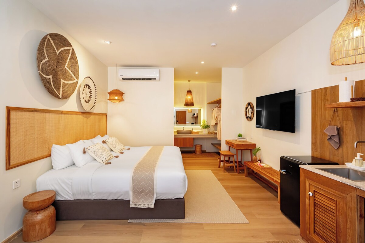 Una habitación de hotel moderna con una cama grande, una kitchenette pequeña, un televisor y un baño visible al fondo.