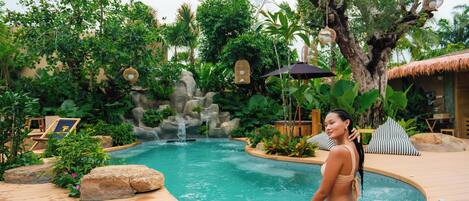 A pool area with a person sitting by the pool, surrounded by tropical plants and a waterfall feature.