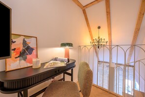 Ein Zimmer mit Holzbalkendecke, einem Tisch mit Lampe, einem Stuhl und einem gerahmten Bild.