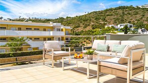 A terrace with outdoor furniture, a table with fruit, and a view of buildings and hills.