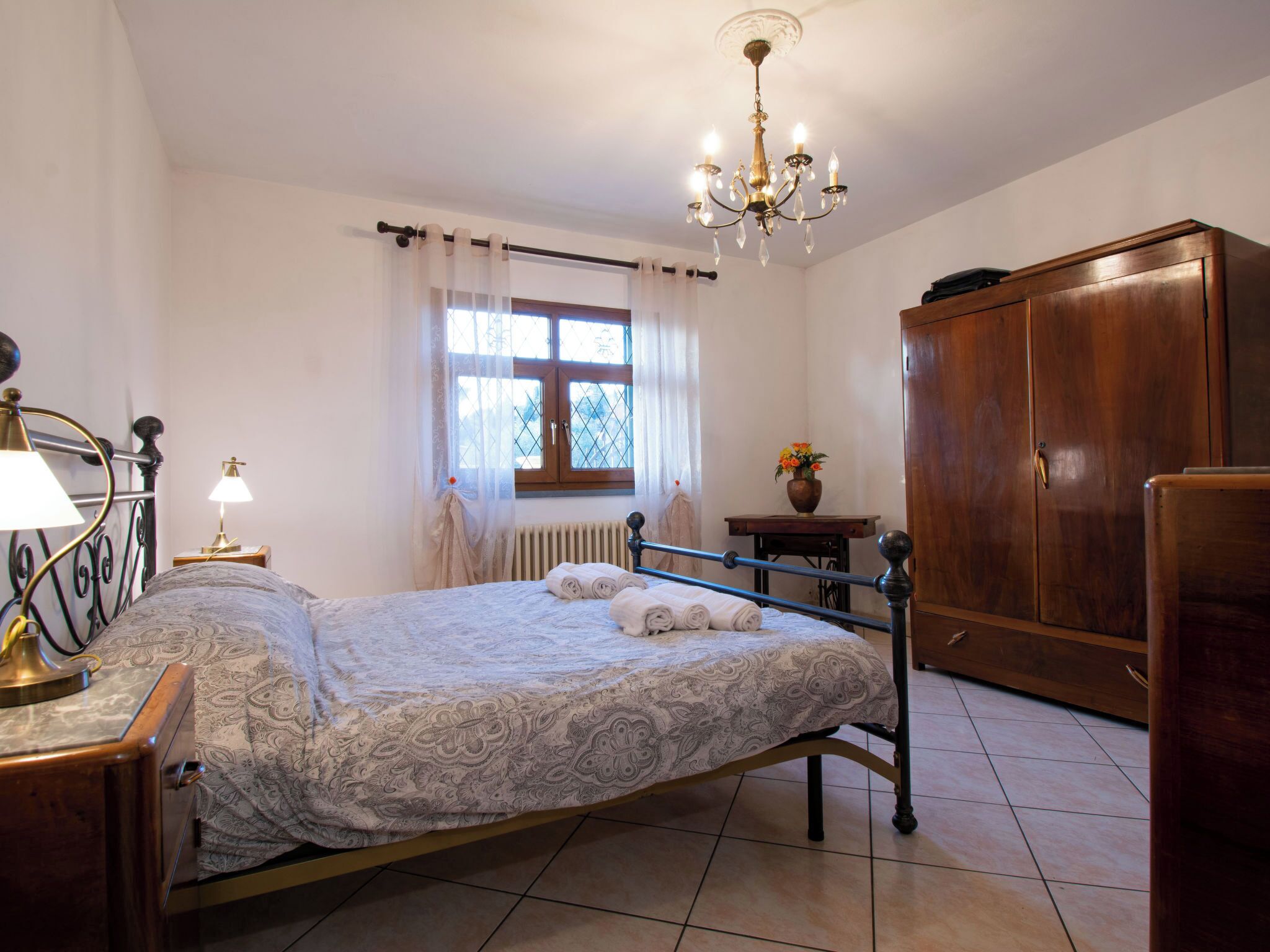 A bedroom with a bed, a wooden wardrobe, a desk with a lamp, a window with curtains, and a chandelier.