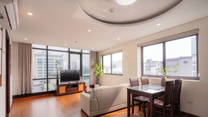 A modern living room with a sofa, TV, dining table, and chairs. Large windows offer a city view. - Sakamoto 1 Apartments by XÔI Residences (Hanoi)