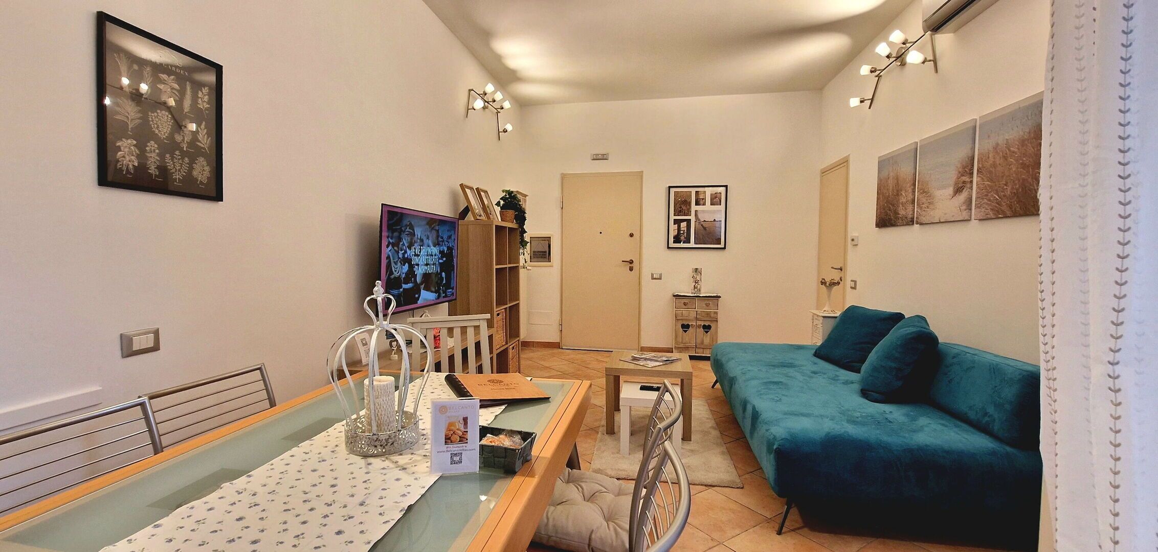 A living room with a teal sofa, a glass coffee table, a television, and a bookshelf.