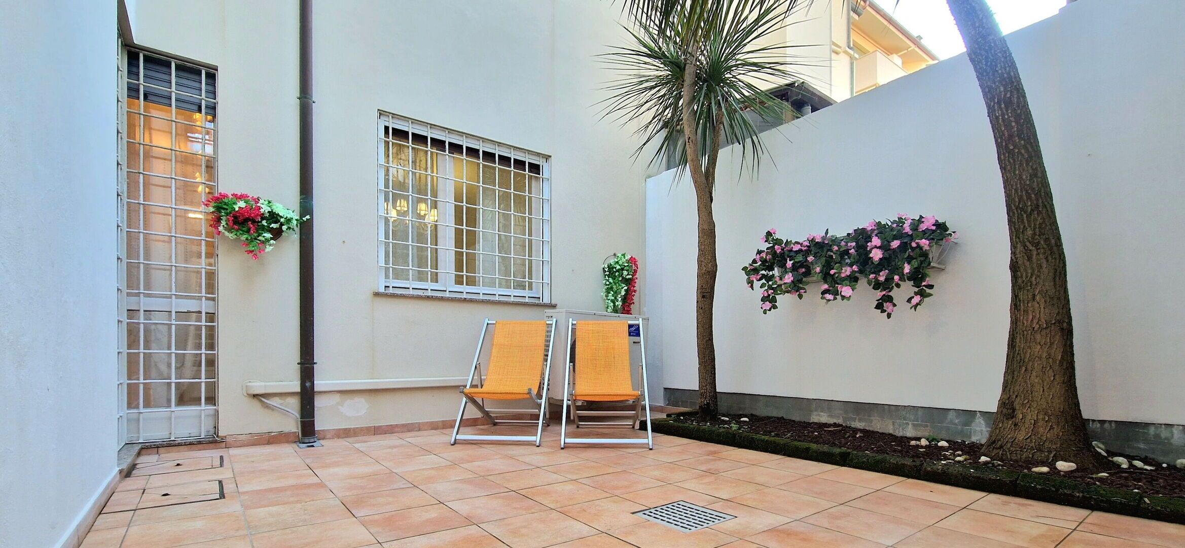 A patio with two chairs, a small table, a window with a metal grill, and a wall with climbing plants.