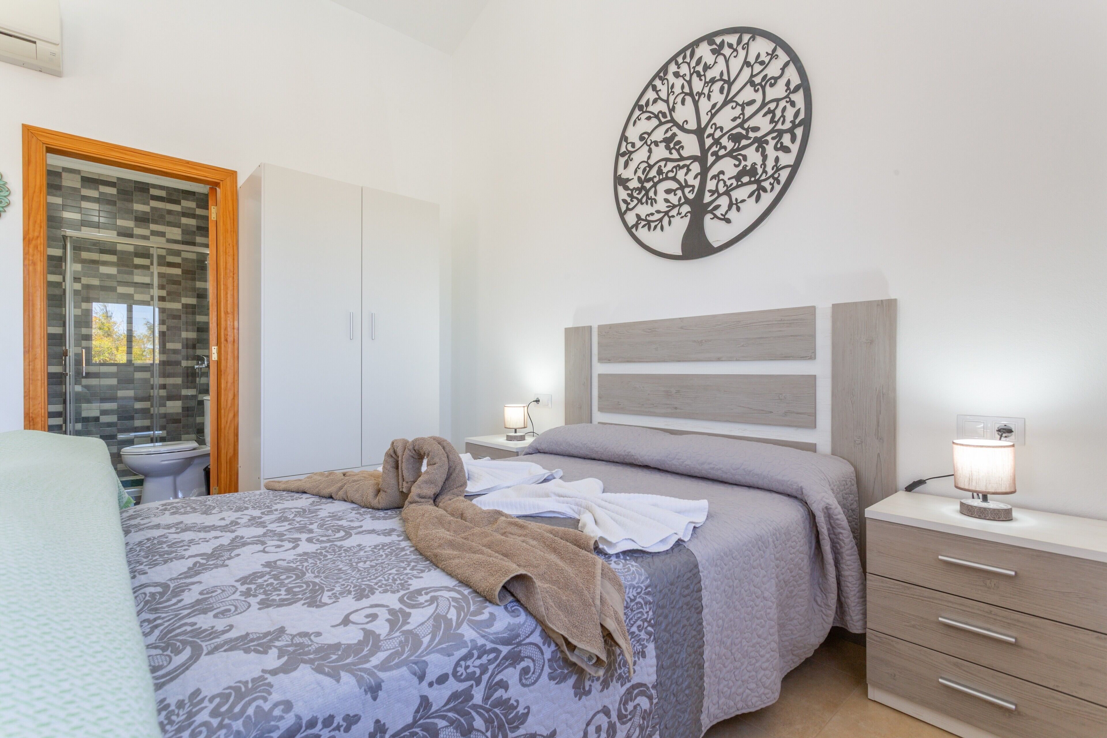 Un dormitorio con cama, mesita de noche, decoración de árbol en la pared, armario de madera y baño con puerta de cristal.