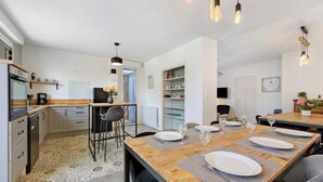 A modern kitchen with a dining area, featuring a wooden table set for four, a refrigerator, and a coffee machine. - La maison du sabotier – Spacious Family Home for 14 Guests (Saint-Malô-du-Bois)