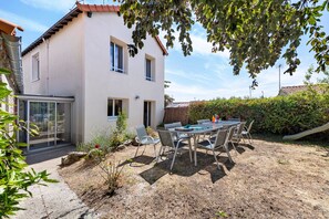 Ett tvåvåningshus med en uteplats som har ett bord och stolar, en liten trädgård och utsikt över omgivningen.