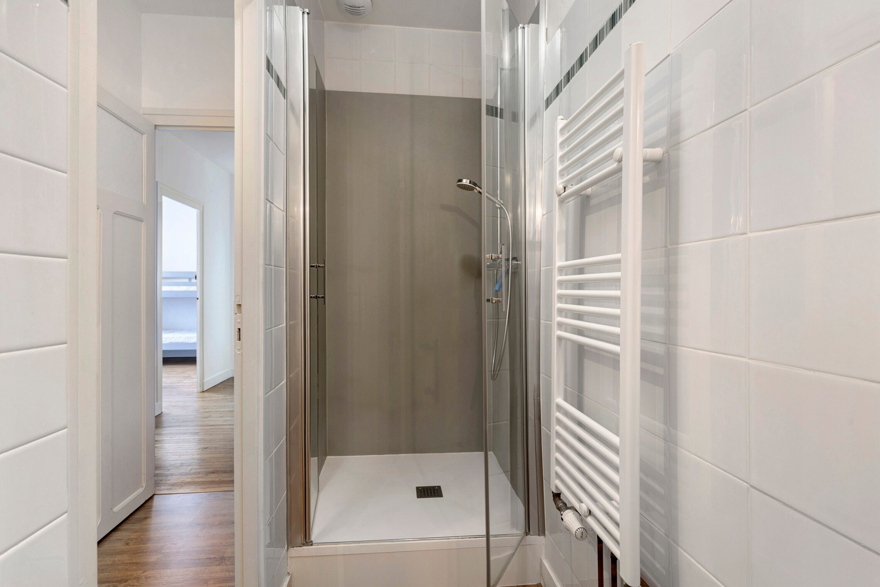 Une salle de bain moderne avec une douche à parois de verre, des murs recouverts de carreaux blancs et un radiateur apparent.