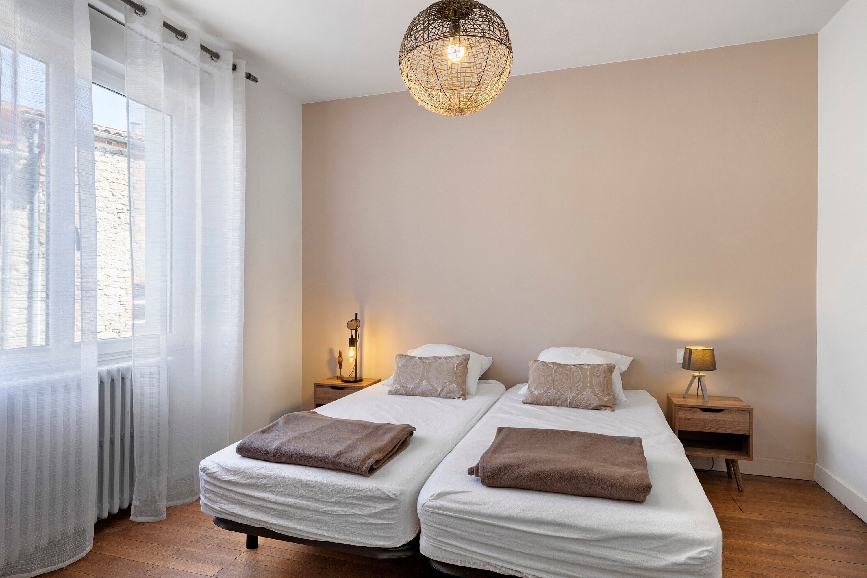 Una cama doble con dos almohadas y una colcha, una mesita de noche con una lámpara, y una ventana con cortinas translúcidas.