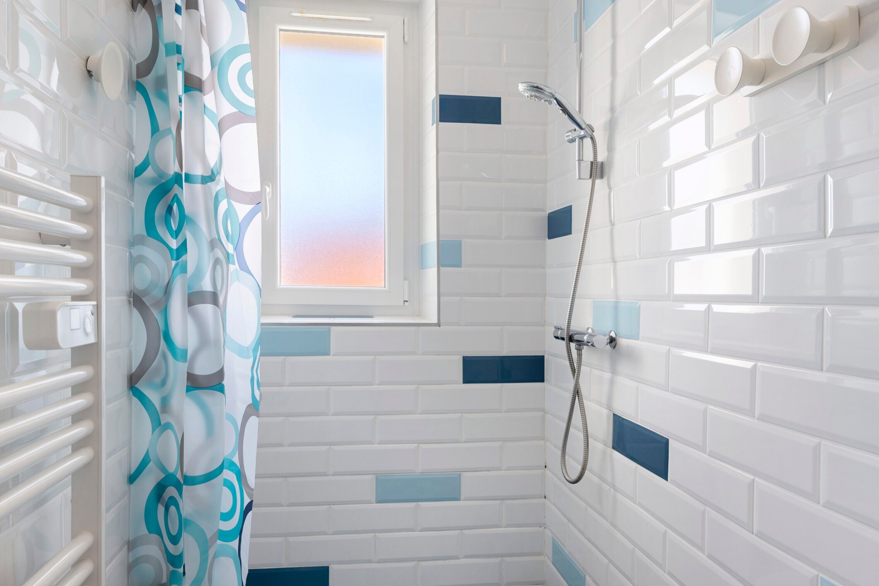Ein Badezimmer mit weiß gefliesten Wänden, einer Dusche mit Duschhandstück und einem Fenster mit Blick.
