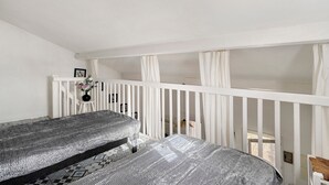 A bedroom with two beds, white railings, a small table with a vase, and a view of the outside.