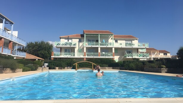 A swimming pool with a clear blue sky, a multi-story building with balconies, and a stone bridge leading to the pool.