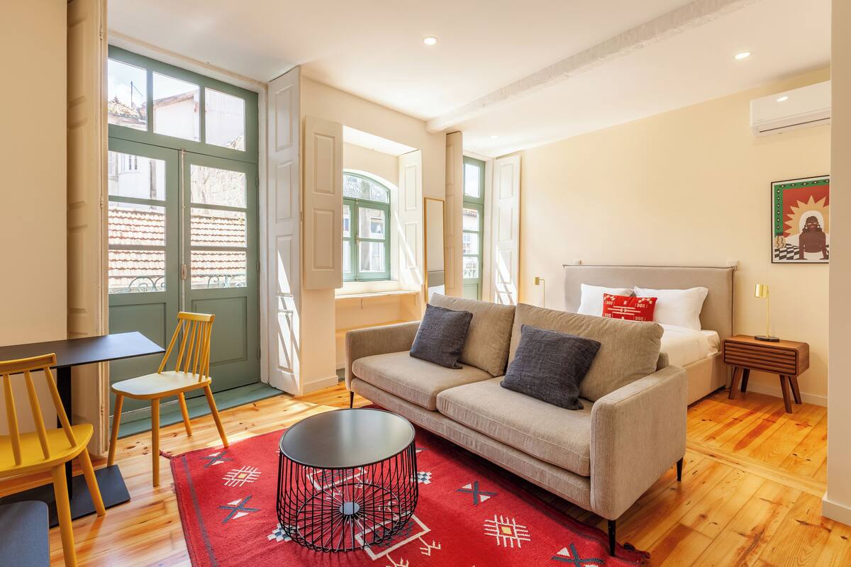A cozy living room with a sofa, a small table, and a chair. There is a bed with a headboard and a nightstand with a lamp.
