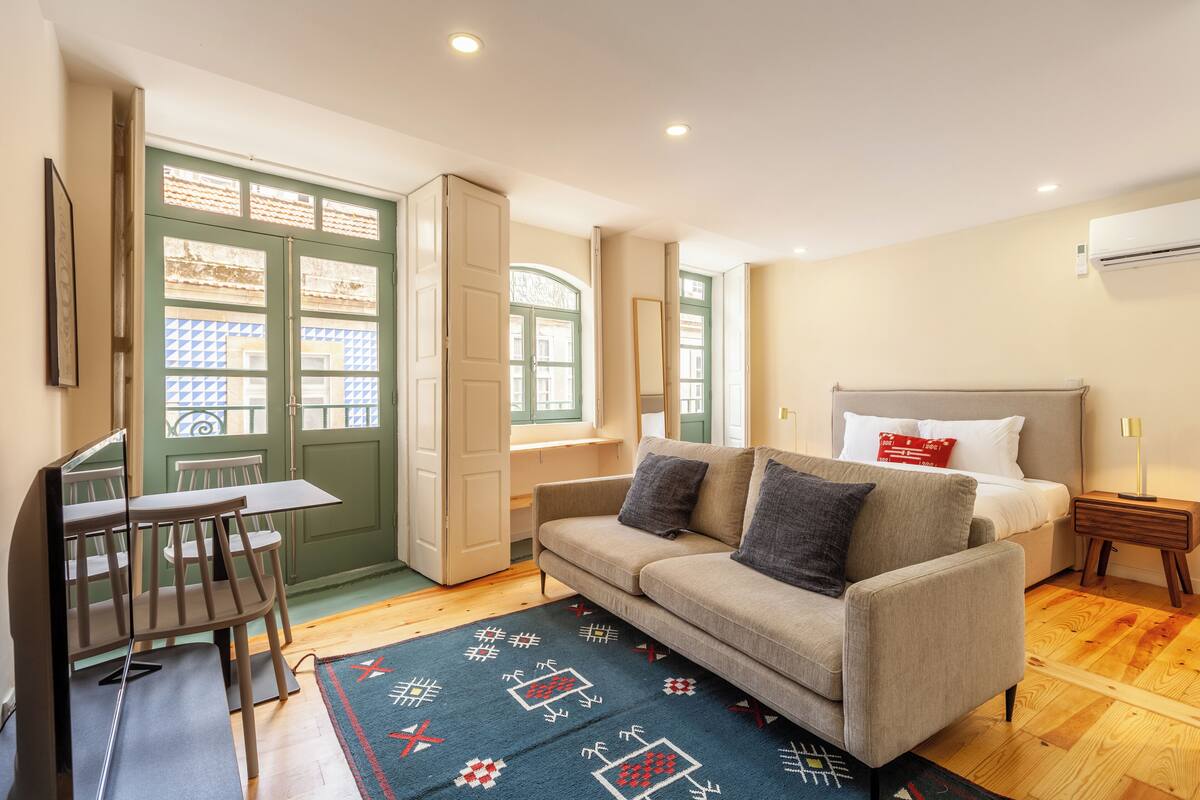 A room with a sofa, a small table, a chair, a bed, a nightstand, and a window with a view of buildings.