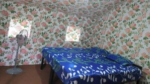 Une pièce avec un papier peint à motifs floraux, un lit recouvert d’une courtepointe bleue et blanche, un ventilateur sur pied et une fenêtre laissant entrer la lumière naturelle.