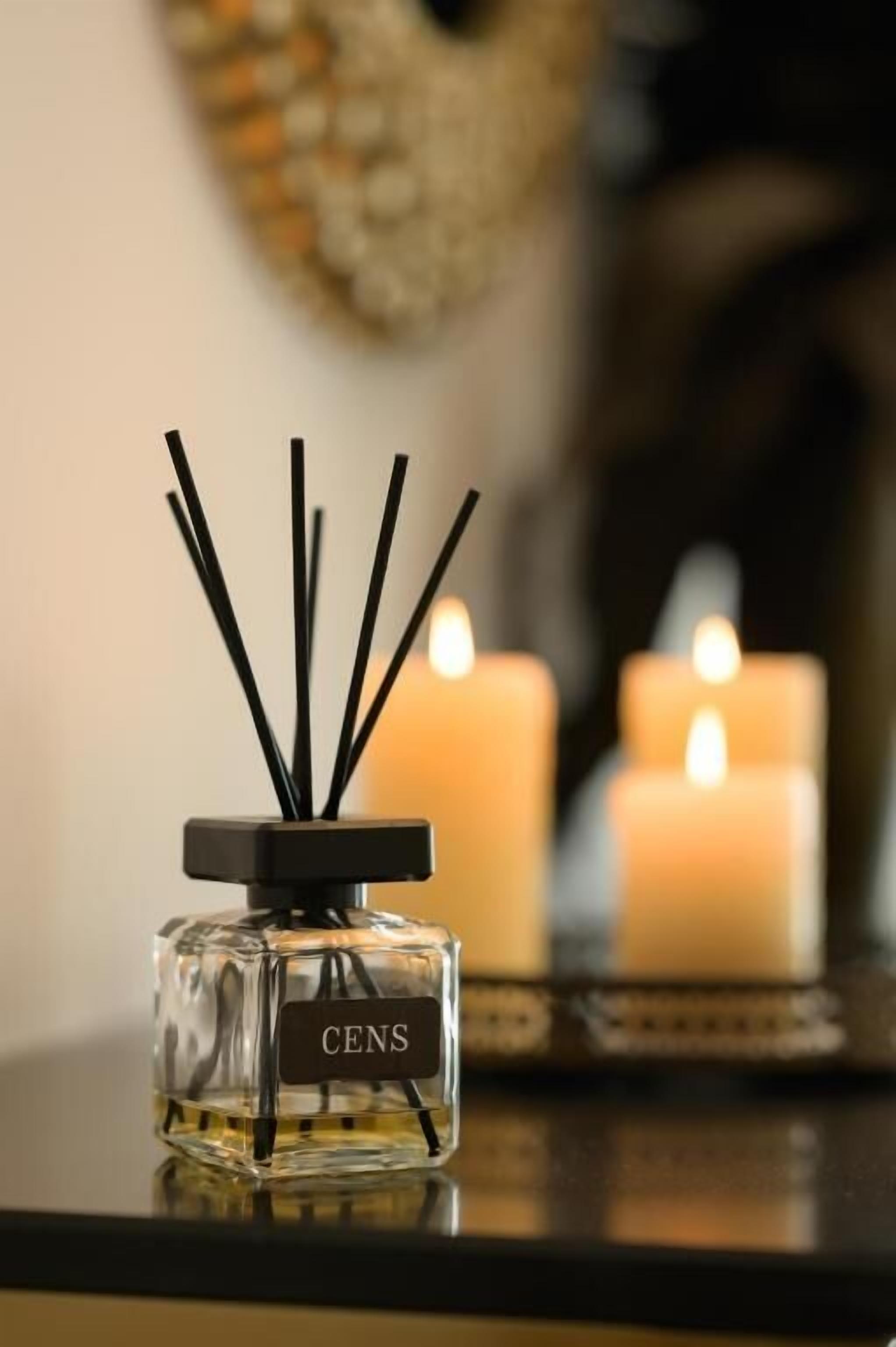 Una botella de fragancia CENS con palitos de fibra en una superficie reflectante, acompañada de dos velas encendidas al fondo.