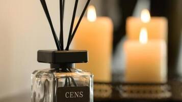 Una botella de fragancia CENS con palitos de fibra en una superficie reflectante, acompañada de dos velas encendidas al fondo.