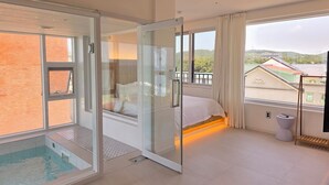 Una habitación de hotel moderna con un jacuzzi de vidrio, una cama con ropa blanca y vistas a un paisaje verde a través de amplios ventanales.
