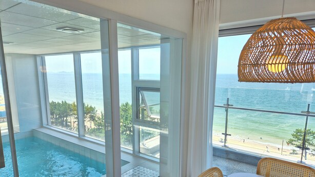 Habitación con un ventanal grande que da a una piscina y al mar. Se puede ver una silla de mimbre y una mesa con mantel blanco.