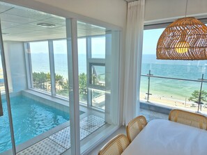 A room with a large window overlooking a pool and the sea. There is a wicker chair and a table with a white tablecloth.