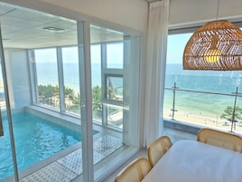A room with a large window overlooking a pool and the sea. There is a wicker chair and a table with a white tablecloth.