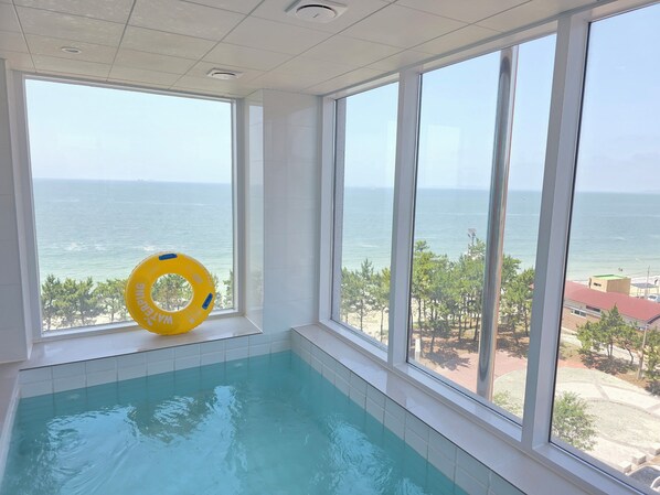 A swimming pool with clear blue water, large windows offering a view of the sea and beach, and a yellow inflatable ring on the pool edge.